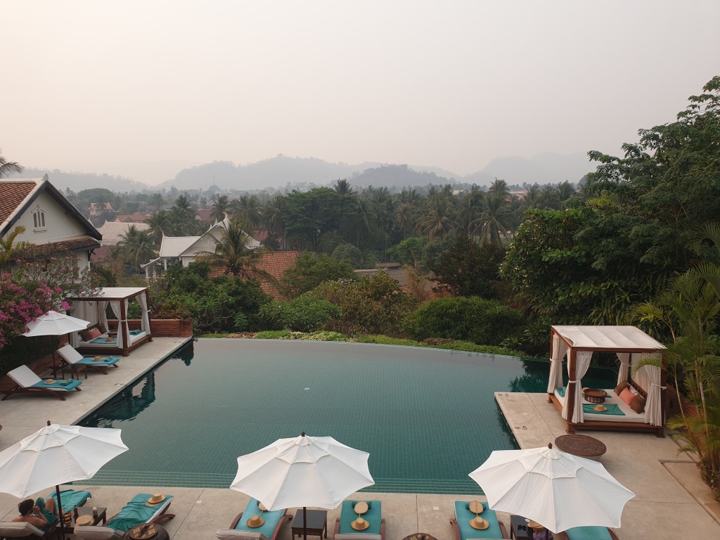 View of a luxurious pool area at a resort in Luang Prabang, surrounded by tropical greenery and mountains in the background, under a hazy sky.