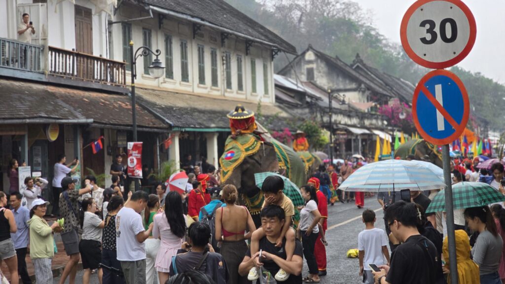 A bustling street scene during the Pi Mai Lao festival in Luang Prabang, featuring elaborately decorated elephants parading amidst a crowd of onlookers, umbrellas, and traditional decorations.