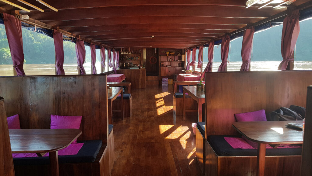 Interior of a riverboat with wooden seating, large windows, and purple curtains, showcasing a relaxed atmosphere for river travel in Laos.